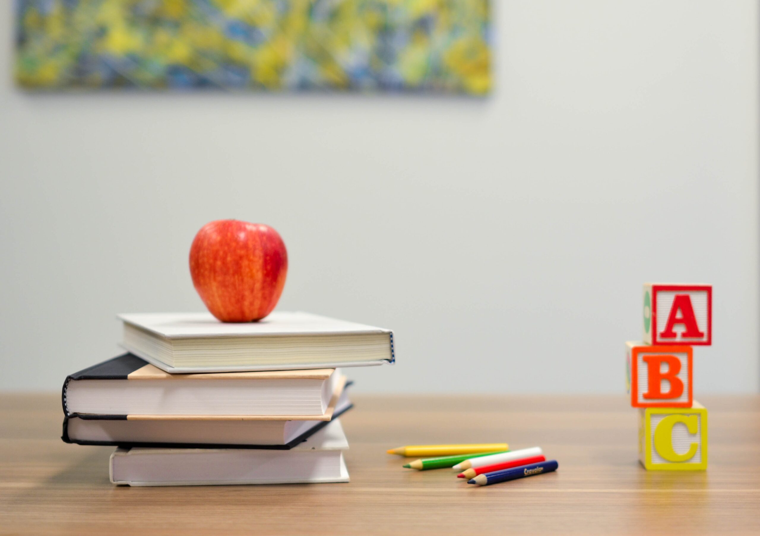 An apple sat on top of a pile of books next to some coloured pencils and 3 blocks spelling out ABC on them vertically